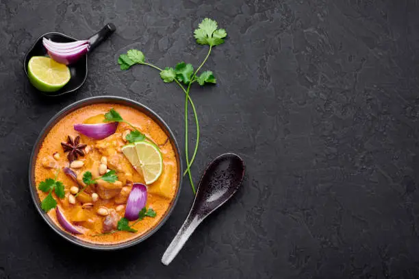 Photo of A Chicken Massaman Curry in black bowl at dark slate background. Massaman Curry is Thai Cuisine dish with chicken meat, potato, onion and many spices. Thai Food. Copy space. Top view