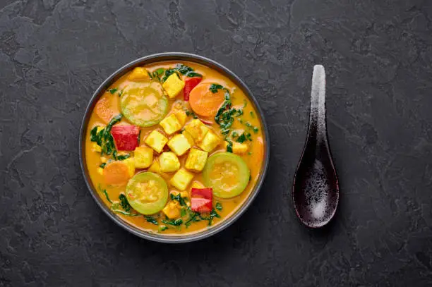 Photo of A Yellow Veg Thai Curry with Tofu and vegetables in black bowl at dark slate background. Vegetarian Thai Curry with tofu, zucchini, bell pepper, spinach, carrot. Thai Food. Indian vegetable curry