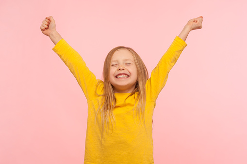 Child sincerely rejoicing success. Cute energetic enthusiastic little girl raising arms in excitement and keeping eyes closed, enjoying victory, winning. indoor studio shot isolated on pink background