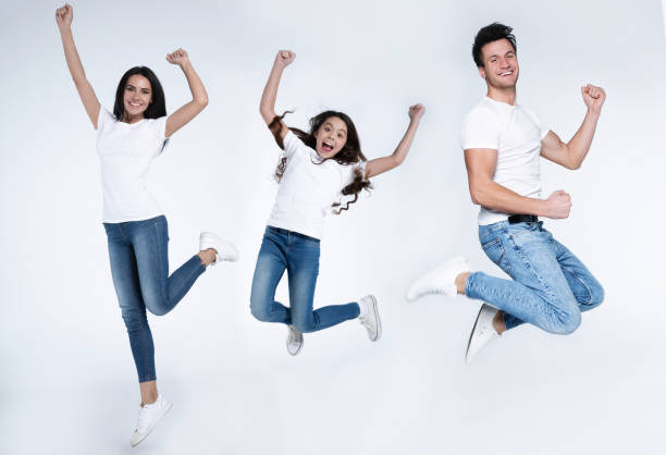 興奮し、幸せな若い美しい家族は、白い背景に孤立した白いtシャツとジーンズでジャンプして叫んでいます。 - couple indoors studio shot horizontal ストックフォトと画像