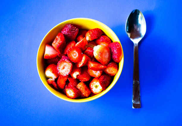 Slices of strawberry in a yellow cup and a spoon on a blue table
