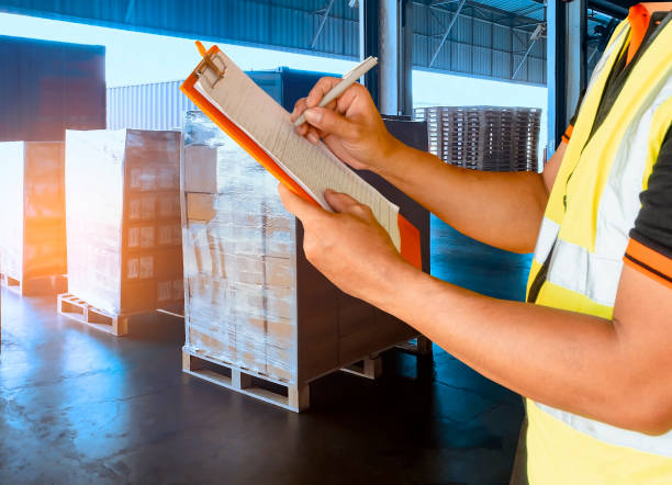 istock Warehouse inventory management. courier shipment transportation. Stack of cardboard boxes on wooden pallet. warehouse worker is writing on clipboard with inspecting the shipment. 1215510050