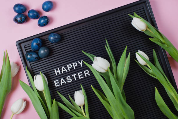 istock Happy Easter - text on black letter board with white tulips and blue colored eggs on pink background. 1215511254