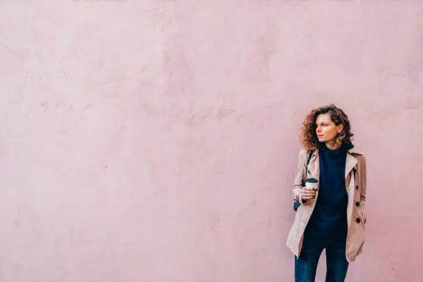 Beautiful young woman with curly hair wearing beige jacket drinking coffee standing near blank pink wall looking away, copy space.