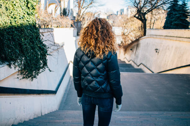 Rear view young woman with long curly hair stands at the top of staircase in city on sunny spring day.