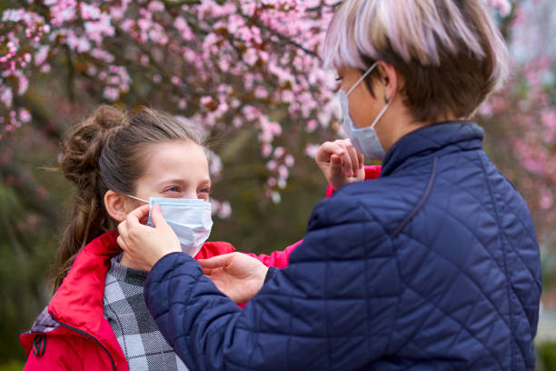 madre e hija con una máscara facial están en la ciudad al aire libre, árboles en flor, temporada de primavera, tiempo de floración - concepto de alergias y protección de la salud contra el aire polvoriento - cherry blossom flower head spring flower fotografías e imágenes de stock