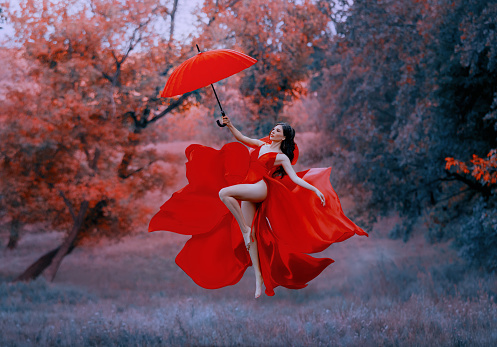 Beautiful elegant happy woman fly with umbrella. Red long sexy silk dress fabric fluttering in wind. goddess dancing joyful cheerful in air. Mystic autumn trees forest. Art fantasy levitation princess