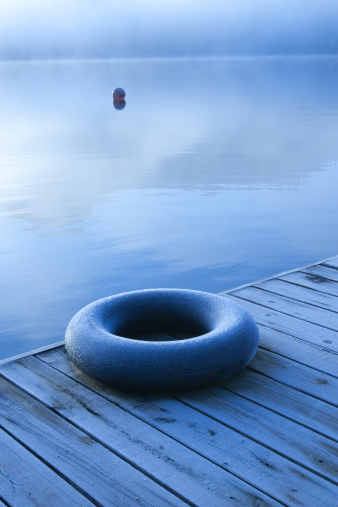 A lone frost-covered inner tube sits on a dock in winter with a buoy and misty lake in background. Overall blue color.