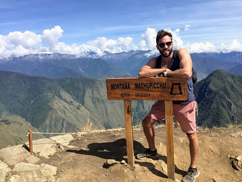 A young male tourist standing beside the sign for the summit of Machu Picchu mountain at the famous Machu Picchu site outside of Cusco, Peru.