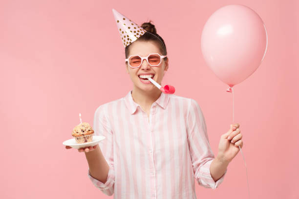 счастливый birhday девушка празднует, держа воздушный шар и торт, дует свисток, изолированные на пастельных розовый фон студии - party hat birthday celebration party стоковые фото и изображения