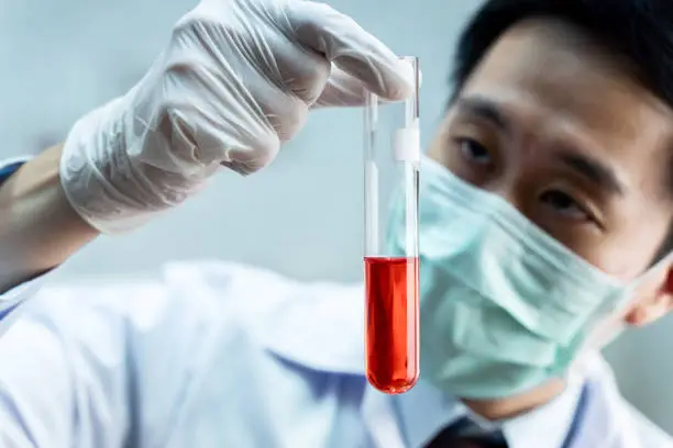 Photo of Close-up of Asian male scientist in white uniform holding laboratory flask with red chemical solution. Chemistry, Science, Biological, Toxicology, Research and Development concept