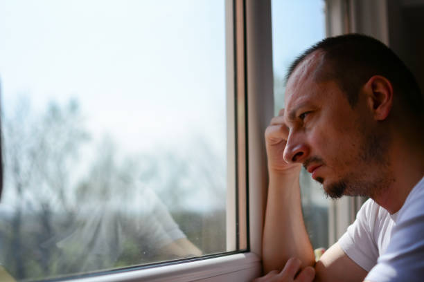 hombre que sufre de depresión mirando por la ventana - salud mental fotos fotografías e imágenes de stock