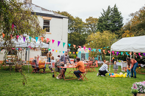 A large group of people are sat enjoying afternoon tea in the garden. Children play with balloons and canopies  are draped with colorful bunting.