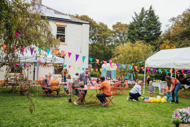 Garden Fun A large group of people are sat enjoying afternoon tea in the garden. Children play with balloons and canopies  are draped with colorful bunting. backyard party stock pictures, royalty-free photos & images