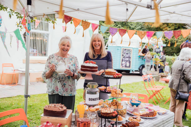 The Garden Fete Two senior ladies stand in a garden decorated in colorful bunting. One holding a pot of tea the other lady a homemade cake covered in strawberries and resting on a cake stand. A table covered in cakes and sweet food stands in front of them and people stand around a pretty painted food truck in the background. fete stock pictures, royalty-free photos & images