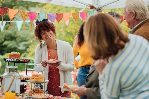 A young mixed race female stands at a table filled with cakes in a garden enjoying afternoon tea whilst chatting to friends. There is colorful bunting hanging from above.