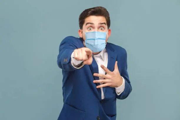 Shocked man with surgical medical mask pointing finger at camera with shocked face. Business people medicine and health care concept. Indoor, studio shot on blue background