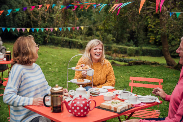 Having A Laugh Adults sat in the garden at a picnic table laughing and enjoying afternoon tea. Colorful bunting blows in the breeze afternoon tea photos stock pictures, royalty-free photos & images