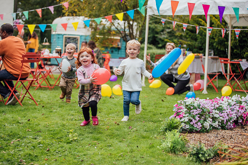 Three young children enjoy playing with balloons at a garden party as they run towards the camera. Adults are sat talking in the background and looking at the children. There is a food truck in the background and a marquee and colorful bunting fill the garden.