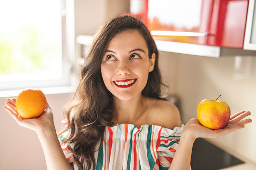 Woman holding apple and orange and standing in her red kitchen