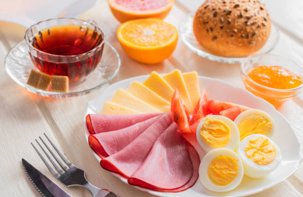 istock Fresh delicious breakfast with soft boiled egg, cheese, ham fresh bread and cup coffee or tea on light wooden background. 1215549266