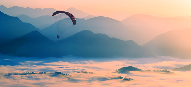 Paraglide silhouette flying over the misty mountain valley in a light of sunrise.