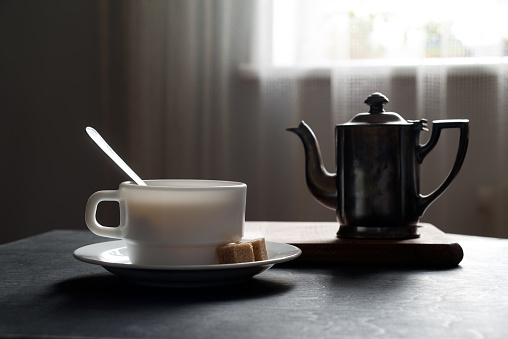 White mug at tea saucer and sugar on black wooden table, stand with a kettle, back lit, curtain