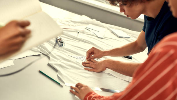 istock Production process. Male hands working with sketches or patterns at studio and preparing fabric. Group of millennials creating clothes together 1215557826