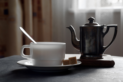 White mug at tea saucer and sugar on black wooden table, stand with a kettle, back lit, curtain