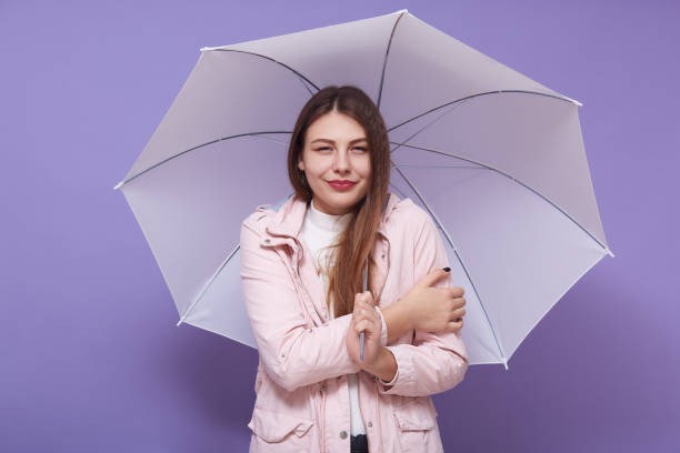 istock Delighted good looking long haired cute female looking directly at camera, cold temperature, holding pink umbrella, standing isolated over lilac bakground in studio. People and weather concept. 1215558760