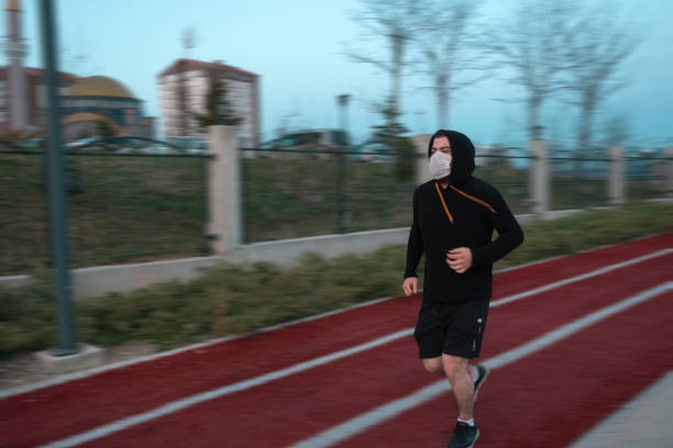 Young man wearing mask and a hoodie running in at a park stock photo