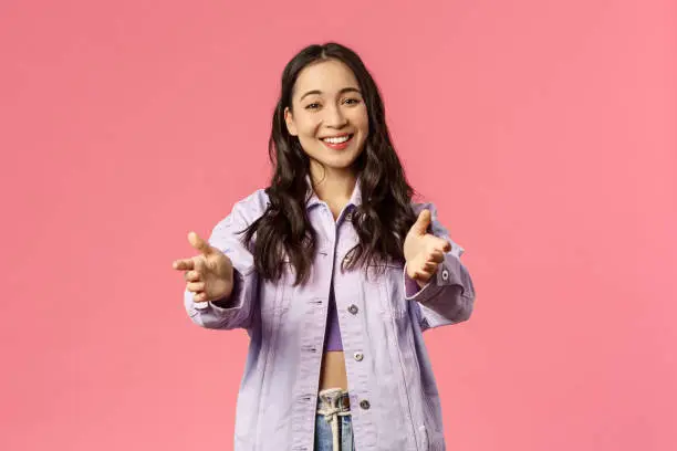 Portrait of cheerful, lovely mixed-raise girl in denim jacket, streetstyle outfit, reaching hands to camera, want hold or hug somone, looking at adorable thing, smiling joyfully, pink background.
