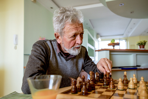 A senior bearded man is playing a chess game at home with his wife. Close-up view of an older man who is making a chess move with a chess piece.