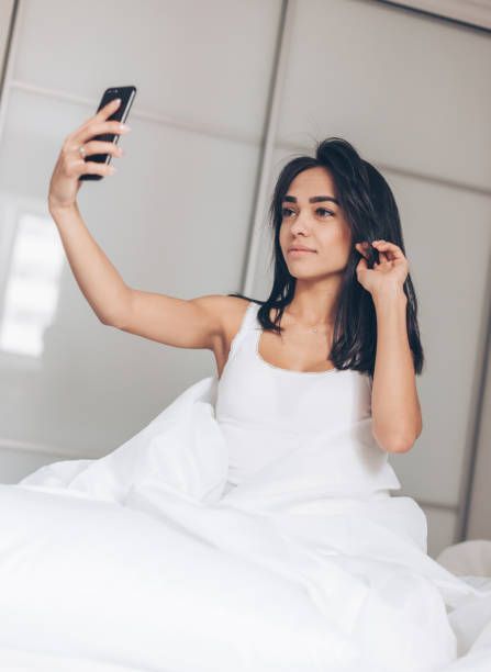 istock Cute young smiling woman sitting on the bed and make a selfie 1215580939