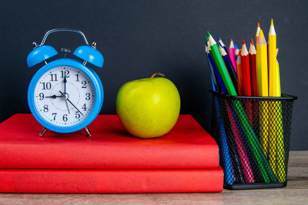istock The blue alarm clock along with multi-colored pencils stand on a stack of red books, next to an apple 1215585724