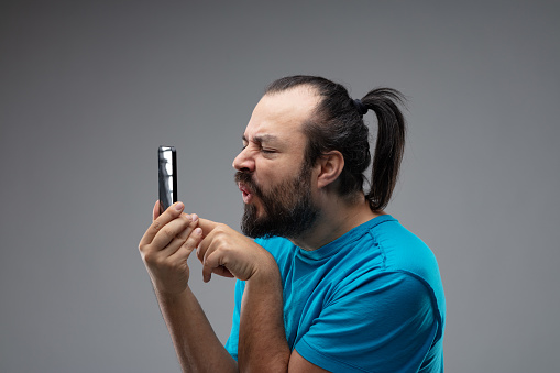Bearded man in blue t-shirt, with black hair combed in ponytail, grimacing and squinting while using smartphone, holding it up close to his face. Side portrait isolated on grey studio background