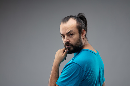 Bearded man with black hair combed in ponytail, standing in blue t-shirt against grey background and looking back at camera with a finger to his chin and calm questioning face. Copy space