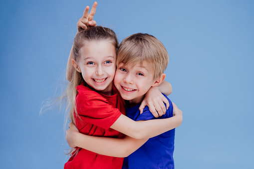 Happy siblings embracing and looking at camera isolated on blue background, in studio