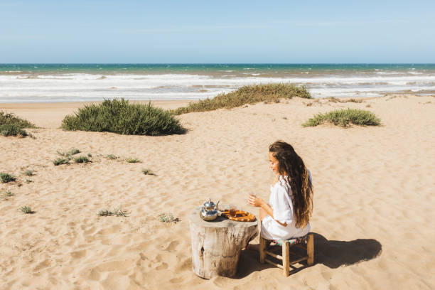 jeune femme heureuse appréciant le thé marocain traditionnel de menthe dehors sur la plage de sable d’océan au maroc. vue par derrière. - at the beach photos et images de collection