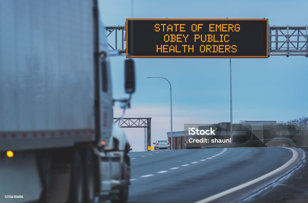 Trucking during State of Emergency State of Emergency Obey Public Health Orders on an overhead highway sign during the Coronavirus pandemic, out of focus semi truck in foreground. Road Sign Stock Photo