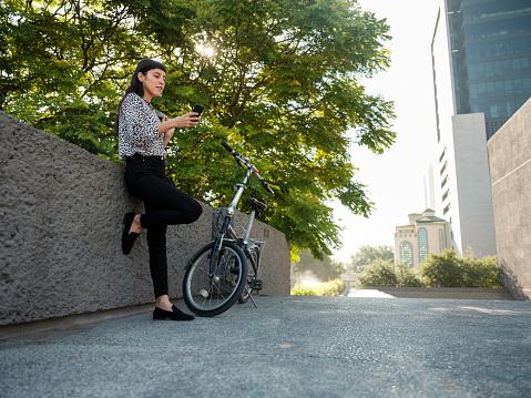 A young mexican woman standing and leaning on a wall outside in the cite, taking a break from her bike and using her phone.