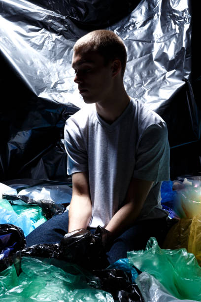 istock Young man seated on plastic bag garbage, environmental - consciousness concept, shoot inside in studio, dark light background. 1215634672