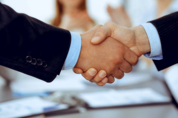 istock Business people shaking hands at meeting while theirs colleagues clapping and applauding. Group of unknown businessmen and women in modern white office. Success teamwork, partnership and handshake concept 1215635744
