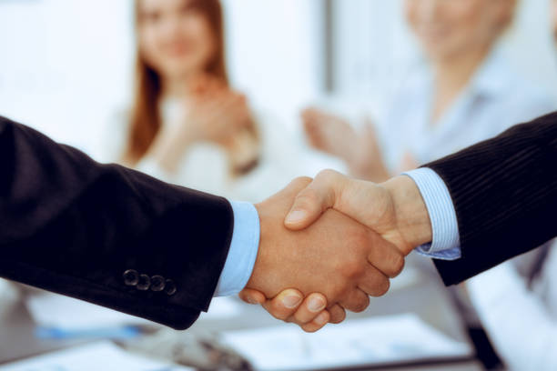 istock Business people shaking hands at meeting while theirs colleagues clapping and applauding. Group of unknown businessmen and women in modern white office. Success teamwork, partnership and handshake concept 1215635754