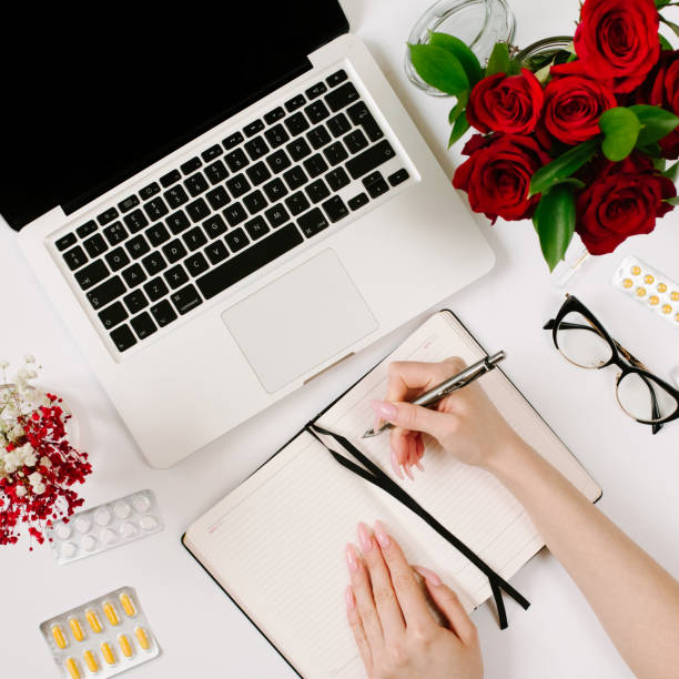 istock Woman hands write in notebook. Work at home when sick. Remote work, epidemic and isolation concept with blister pills, roses bouquet, laptop and notebook 1215643414