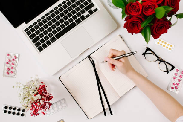 istock Woman hands write in notebook. Work at home when sick. Remote work, epidemic and isolation concept with blister pills, roses bouquet, laptop and notebook 1215643425