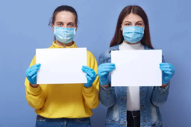 Photo of Image of two young women wearing medical disposable masks and medical gloves holding white blank papers in hands, copy space for some inscriptions. Corona virus, covid 19, health care concept.