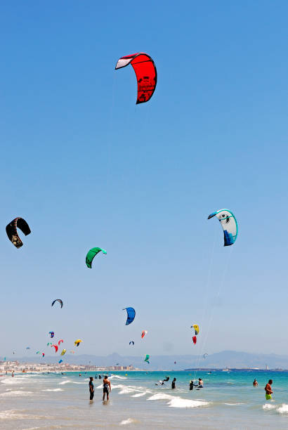 People practising KiteSurfing at Tarifa beach in South Spain People practising KiteSurfing at Tarifa Beach in Spain on a sunny summer day. Tarifa is pretty famous in the kitesurfing world, probably known best for its strong wind. Because of its location at the very tail end of Spain, Tarifa sits 15 kilometers away from Morocco in North Africa. With the Strait of Gibraltar in between, a wind funnel is created between the two land masses which means Tarifa enjoys strong winds all year round. kite sailing stock pictures, royalty-free photos & images