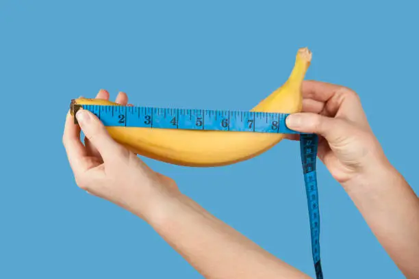 Photo of Measuring the size of a banana as a symbol of the male penis isolated on blue background. Big dick length. Strong erection and impotence problem