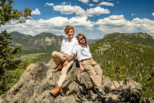 A couple is sitting on a giant rock, leaning to each other and looking at the camera with greenish hills and blue sky as the background.